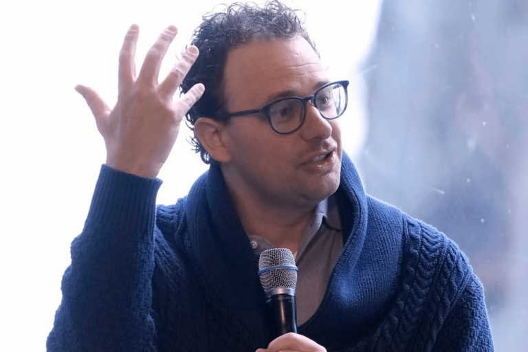 Man wearing glasses and a blue knit cardigan speaks into a handheld microphone while gesturing with his left hand.