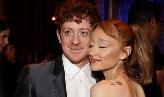 Man in a tuxedo and a woman in a pink dress pose closely at a formal event, smiling gently at the camera.