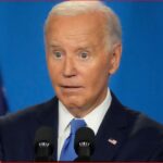 A man with white hair in a dark suit and blue tie speaks at a podium with two microphones, American flags in the background, looking surprised or startled.
