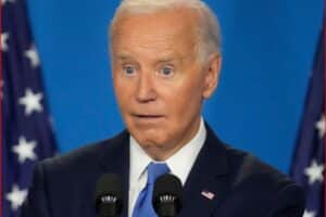 A man with white hair in a dark suit and blue tie speaks at a podium with two microphones, American flags in the background, looking surprised or startled.