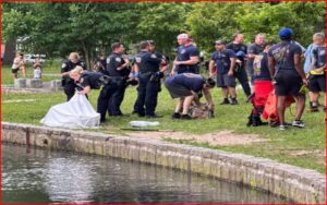Group of uniformed responders and bystanders on a grassy riverbank; a white sheet covers something near the water, indicating a recovery operation.