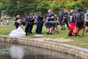 Group of uniformed responders and bystanders on a grassy riverbank; a white sheet covers something near the water, indicating a recovery operation.
