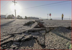 Wide view of cracked asphalt with a large pothole in the foreground; a person in a yellow jacket stands near a stop sign in the distance under power lines.