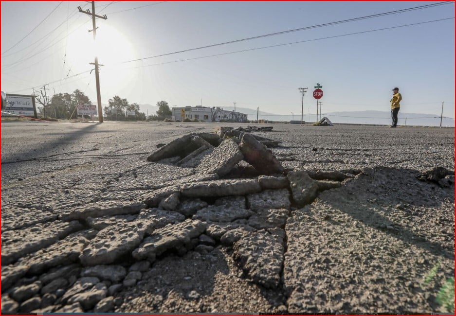 Wide view of cracked asphalt with a large pothole in the foreground; a person in a yellow jacket stands near a stop sign in the distance under power lines.