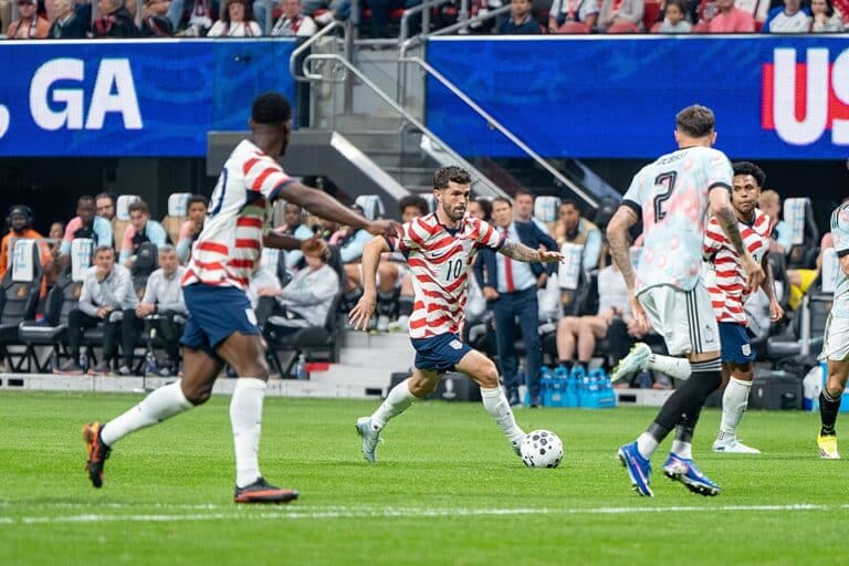 Soccer player in red-and-white striped jersey (number 10) dribbles toward the ball on a green field with teammates nearby and a stadium crowd behind him