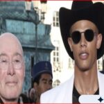 Two men walk outdoors at a busy urban event: one bald with glasses, the other wearing a black wide-brim hat and round sunglasses in a white jacket.