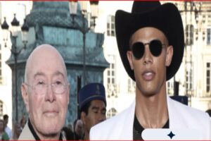 Two men walk outdoors at a busy urban event: one bald with glasses, the other wearing a black wide-brim hat and round sunglasses in a white jacket.