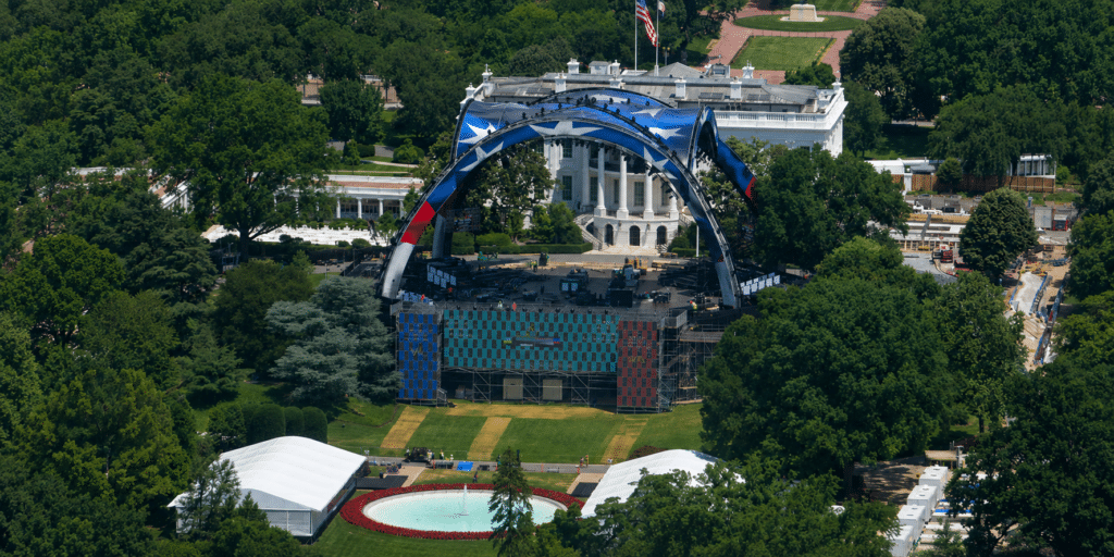 Aerial view of a large outdoor concert stage on the White House lawn, featuring a blue arched structure and colorful video screens amid trees.