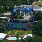 Aerial view of a large outdoor concert stage on the White House lawn, featuring a blue arched structure and colorful video screens amid trees.