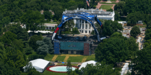 Aerial view of a large outdoor concert stage on the White House lawn, featuring a blue arched structure and colorful video screens amid trees.