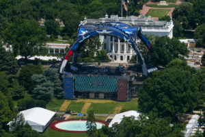 Aerial view of a large outdoor concert stage on the White House lawn, featuring a blue arched structure and colorful video screens amid trees.