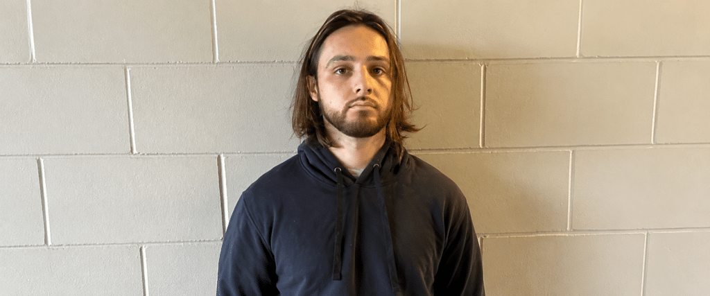 Man with shoulder-length brown hair and a beard, wearing a dark blue hoodie, standing against a gray cinder block wall.