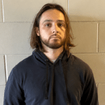 Man with shoulder-length brown hair and a beard, wearing a dark blue hoodie, standing against a gray cinder block wall.