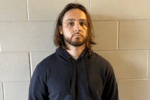 Man with shoulder-length brown hair and a beard, wearing a dark blue hoodie, standing against a gray cinder block wall.
