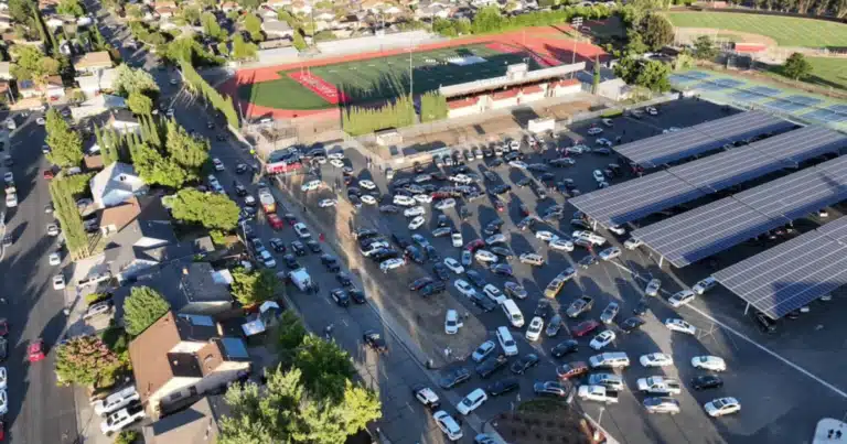 Aerial view of a school campus with a crowded parking lot, a red-run track field, and large solar-panel canopies over parking areas.