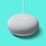 Close-up of a gray fabric-coated spherical smart speaker hanging from a white cable against a bright turquoise background.