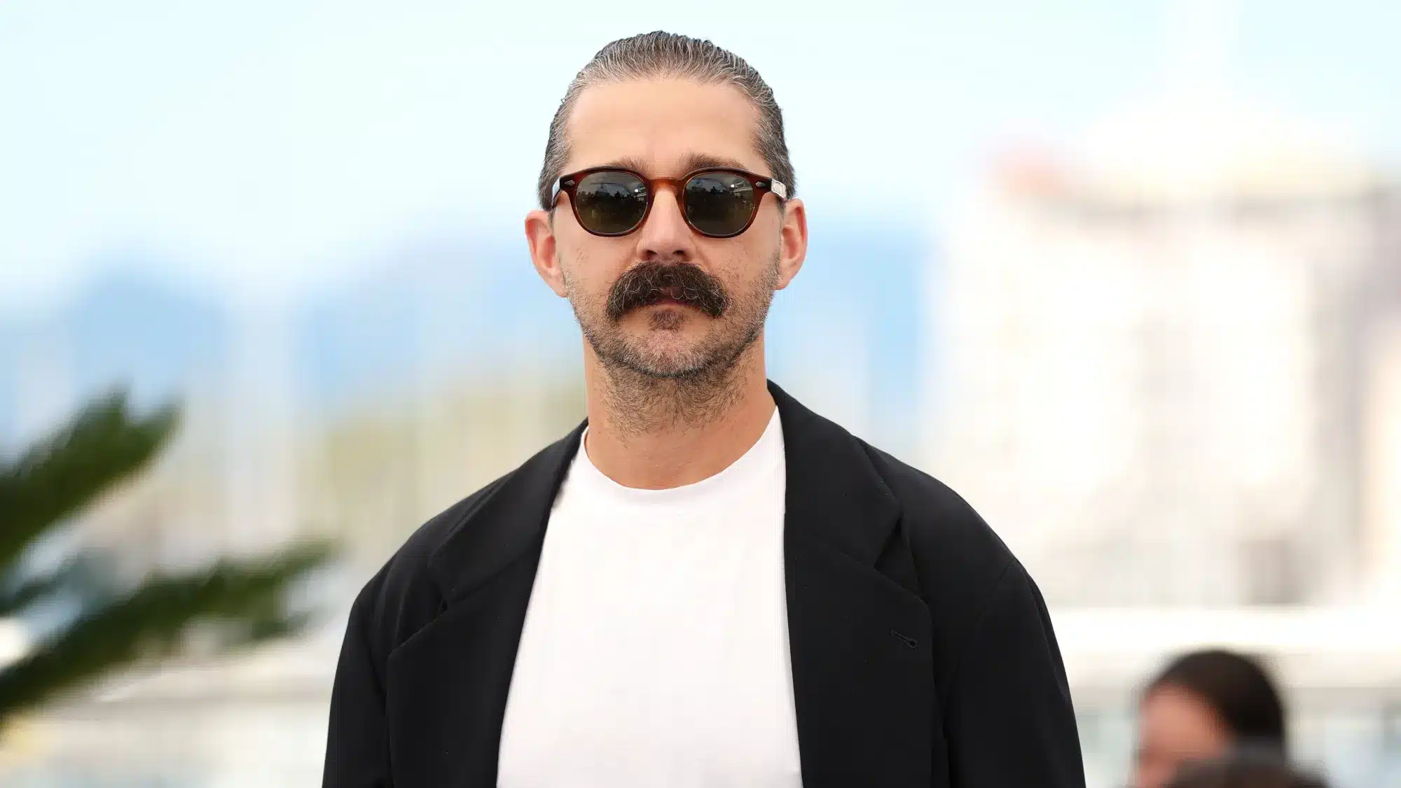 Man with slicked-back hair, thick mustache, and round sunglasses facing the camera, wearing a white shirt and black blazer outdoors.