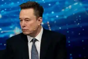 Man in a dark suit and white shirt with a lapel mic, seated on a stage against a blue abstract background from a panel discussion.