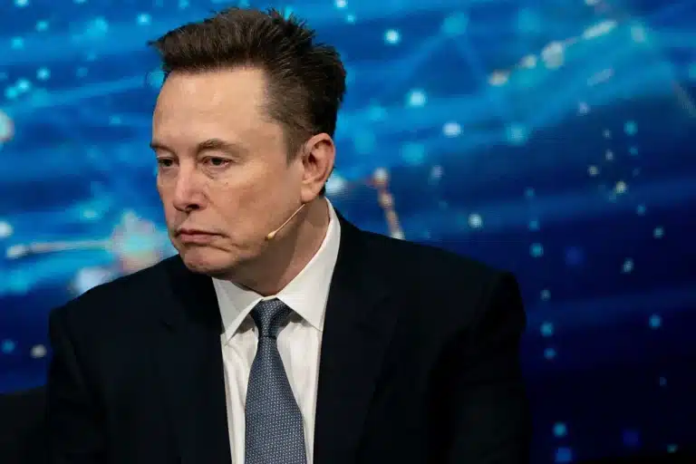 Man in a dark suit and white shirt with a lapel mic, seated on a stage against a blue abstract background from a panel discussion.
