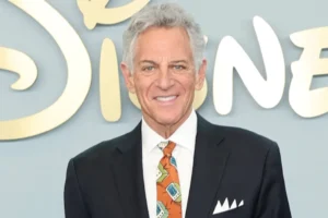 Smiling older man in a black suit and orange patterned tie poses in front of a Disney backdrop.
