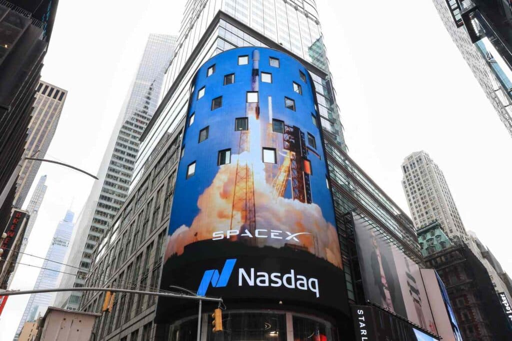Curved blue SpaceX billboard on a Nasdaq building in Times Square showing a rocket launch and the Nasdaq sign below.
