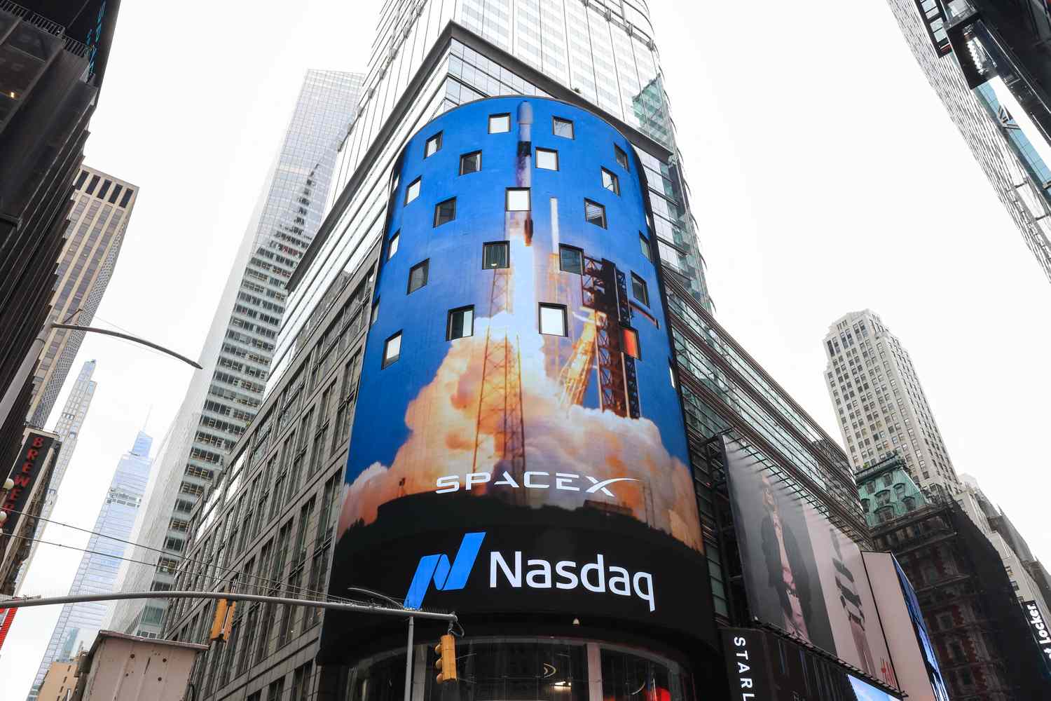 Curved blue SpaceX billboard on a Nasdaq building in Times Square showing a rocket launch and the Nasdaq sign below.