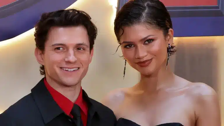 Two people posing together at an event, smiling for the camera; man in a black suit with a red shirt, woman in a black dress with long earrings.