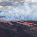 Volcanic lava stream flows across dark terrain with thick white smoke rising above it.