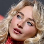 Close-up of a blonde woman with blue eyes and glossy pink lips, wearing makeup and a red sequined top, looking to the side.