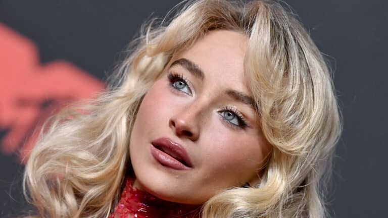 Close-up of a blonde woman with blue eyes and glossy pink lips, wearing makeup and a red sequined top, looking to the side.