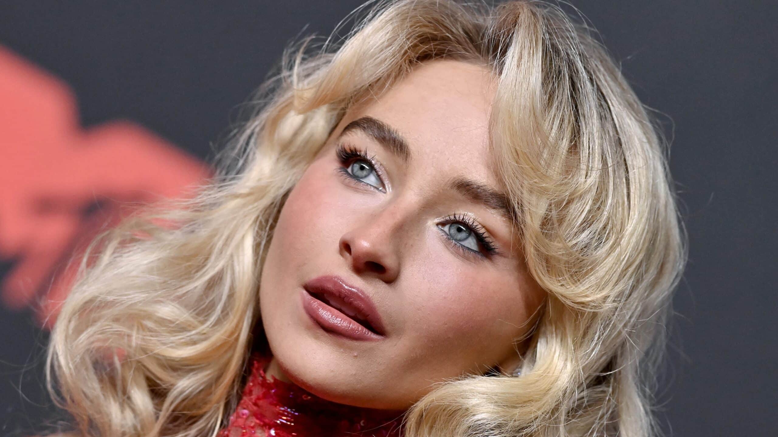 Close-up of a blonde woman with blue eyes and glossy pink lips, wearing makeup and a red sequined top, looking to the side.