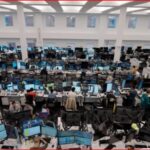 Large open-plan office with rows of desks and dozens of computer monitors, workers at stations.