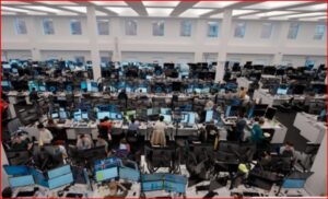 Large open-plan office with rows of desks and dozens of computer monitors, workers at stations.