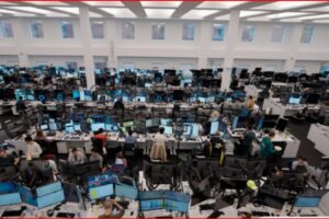 Large open-plan office with rows of desks and dozens of computer monitors, workers at stations.