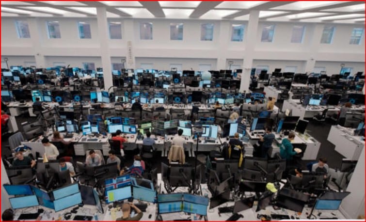 Large open-plan office with rows of desks and dozens of computer monitors, workers at stations.