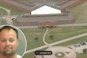 Aerial view of a fenced prison complex with a central building and surrounding grounds; a circular inset shows a man's mugshot in the lower-left corner.