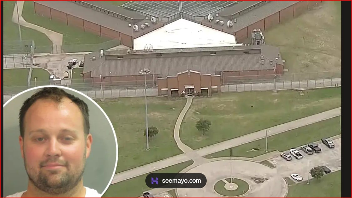 Aerial view of a fenced prison complex with a central building and surrounding grounds; a circular inset shows a man's mugshot in the lower-left corner.