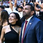Couple in formal attire at a crowded event, smiling as fans and photographers surround them on the red carpet.