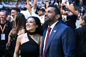 Couple in formal attire at a crowded event, smiling as fans and photographers surround them on the red carpet.