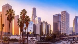 Sunset over a modern city skyline with tall glass buildings, palm trees, and an elevated highway in the foreground.