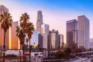 Sunset over a modern city skyline with tall glass buildings, palm trees, and an elevated highway in the foreground.