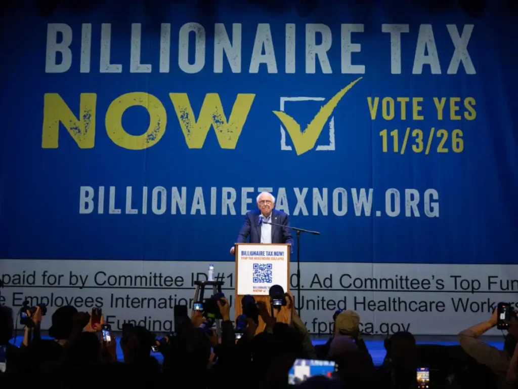 Speaker at a podium in front of a large blue banner that reads 'BILLIONAIRE TAX NOW' with a yellow checkmark and 'VOTE YES 11/3/26'; crowd in foreground taking photos.