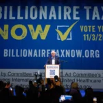 Speaker at a podium in front of a large blue banner that reads 'BILLIONAIRE TAX NOW' with a yellow checkmark and 'VOTE YES 11/3/26'; crowd in foreground taking photos.