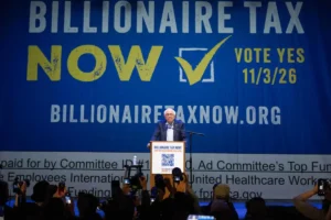 Speaker at a podium in front of a large blue banner that reads 'BILLIONAIRE TAX NOW' with a yellow checkmark and 'VOTE YES 11/3/26'; crowd in foreground taking photos.