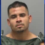 Mugshot of a man with short dark hair and minor facial injuries, wearing a gray jail uniform against a gray backdrop.