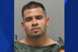 Mugshot of a man with short dark hair and minor facial injuries, wearing a gray jail uniform against a gray backdrop.