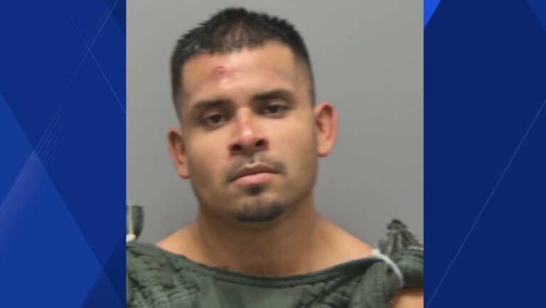 Mugshot of a man with short dark hair and minor facial injuries, wearing a gray jail uniform against a gray backdrop.