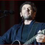 Male musician playing an acoustic guitar on stage, singing into a microphone.