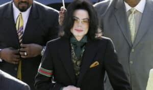 Person in a black suit with a green tie, gold pocket square, patterned vest, and sunglasses, standing outdoors alongside two men in suits.