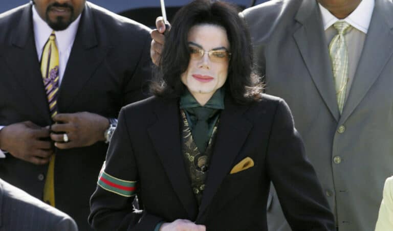 Person in a black suit with a green tie, gold pocket square, patterned vest, and sunglasses, standing outdoors alongside two men in suits.